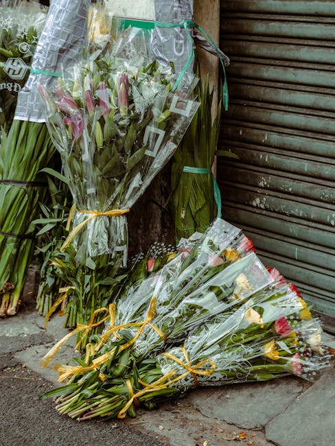 Several bouquets of fresh flowers resting against a wall outside a shop, wrapped in clear plastic and tied with yellow and green ribbons. The arrangements include pink lilies, yellow roses, and variou