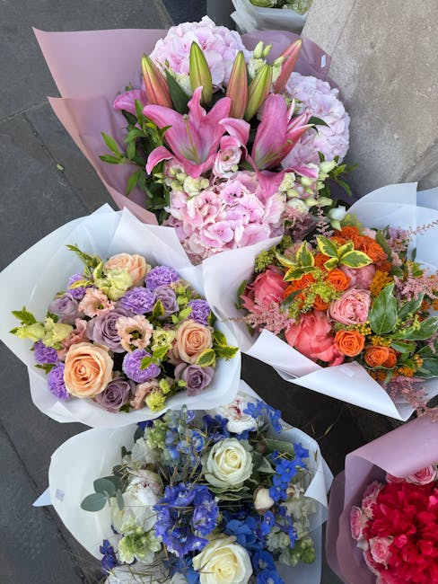 An arrangement of four floral bouquets wrapped in pastel and white paper, placed on a concrete surface. The largest bouquet features pink lilies, pink hydrangeas, and unopened lily buds, with green le