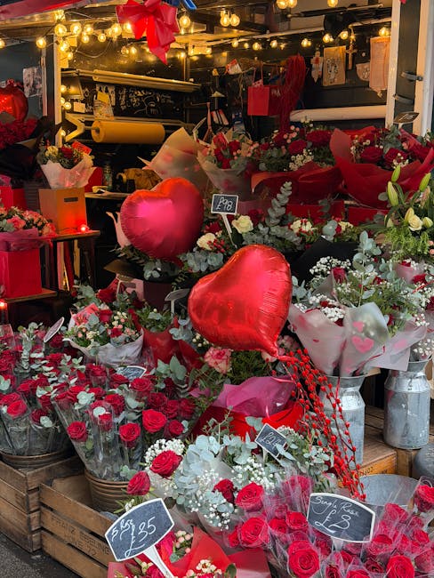 An outdoor floral display showcasing a variety of fresh flowers arranged in bouquets and individual stems, predominantly featuring red roses, white baby's breath, and greenery. Among the arrangements 