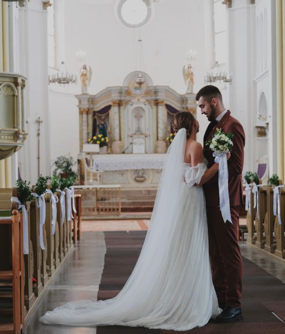 A bride and groom stand close together inside a bright, elegant church, the bride wearing a white, flowing wedding gown with a long train and delicate lace details on her off-the-shoulder sleeves, hol