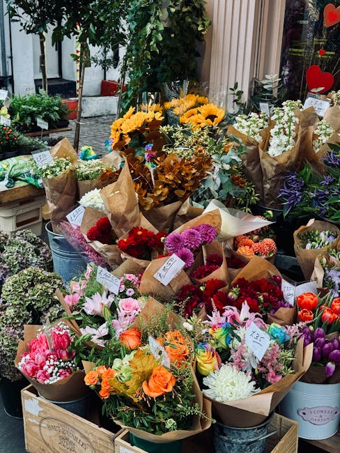 A vibrant display of freshly cut flowers arranged in assorted paper wraps and buckets inside a flower shop. The arrangements include bright yellow sunflowers, pink lilies, red and purple carnations, o