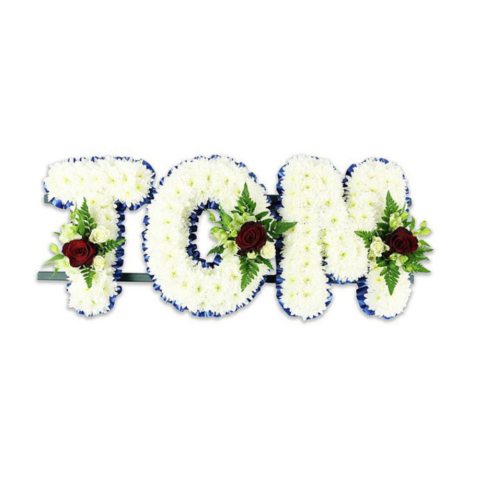 This personalised sympathy display spells 'TOM' in large, sculpted letters made from densely clustered white chrysanthemum blossoms, each tiny flower forming a soft, tactile surface that reads as luminous and pure. The letters are crisply edged in a vivid royal blue ribbon, the narrow border giving each character a refined frame and a cool, elegant contrast to the warm whiteness of the blooms. Within each letter a carefully composed floral spray brings focus: a single, dramatic deep red rose anchors the grouping, its velvety petals suggesting deep respect, while small ivory spray roses and buds soften the effect and provide gentle highlights. Sprigs of fern and lush green leaves thread through these clusters, adding line, texture and a faint, earthy aroma that balances the sweetness of the roses. Beneath, a slender dark green or black metal bar supports the letters, hinting at the practical craftsmanship required to present a name upright at a funeral or memorial. Set against a pure white backdrop and lit to show texture and hue without shadow, the tribute feels composed and solemn-an emblem of remembrance that reads clearly from a distance. Neighbours in Upper Clapton, from Millfields Park visitors to those coming from Lea Bridge Road, often choose this kind of bespoke lettering to honour someone's life; the arrangement's formal lines and tactile softness make it an intimate and public gesture of farewell.