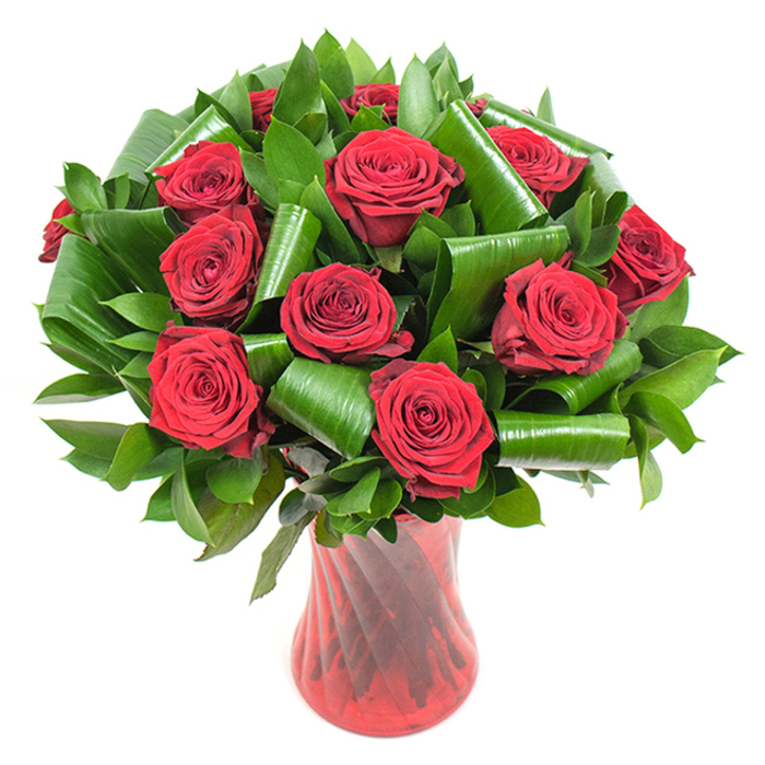 In this image a generous cluster of more than a dozen deep, crimson-red roses forms a luxurious, rounded bouquet, each flower opening into rich, velvety spirals whose petals overlap with a dense, tactile elegance. The blooms are set against an abundant framework of dark green foliage: smaller, glossy leaves that create a lush, supportive background, and broader, bright green leaves that have been folded, curled, and rolled into sculptural cylinders and sweeping loops. Those folded leaves lend the arrangement a contemporary, almost architectural accent, cradling the rose heads and adding depth and movement to an otherwise classic composition. The bouquet is presented in a tall, narrow ruby-red glass vase with a subtle twisted or ribbed texture; the vase's vertical lines pick up ambient light and reflect it back into the crimson tones of the roses, giving the whole piece a quietly dramatic sheen. With a crisp white backdrop and soft, even studio illumination, every petal edge and leaf curl is visible, creating a vivid study in color, texture, and balance. The arrangement suggests warmth and romantic intent-the scent of freshly opened roses and the tactile contrast of smooth glass and velvety petals-and feels at home as an anniversary gift, a Valentine's gesture, or a sophisticated dining-centrepiece. Locally, this thoughtful, refined style would fit perfectly among deliveries for intimate celebrations around Hackney Downs and the quieter terraces leading off Stamford Hill, reflecting the neighbourhood's appreciation for carefully crafted floral design.