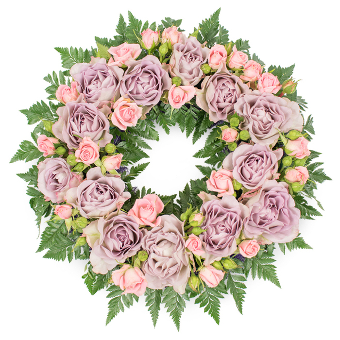 In this overhead image, a perfectly circular funeral wreath lies against a pure white backdrop, giving full focus to its layered flowers and foliage. The ring is lavishly filled with roses, the outer and inner edges softened by petals and fern rather than hard lines. Central to the design are large, fully opened roses in nuanced tones of dusty lavender, soft mauve, and a cool, greyish-purple. Their petals are densely packed and slightly cupped, spiralling inward to form rich, textured centres that create visual depth all around the wreath. Between these main blooms, clusters of delicate blush-pink spray roses thread through the design, their smaller heads and varying stages-from bud to open-bringing a sense of gentle movement and youthfulness. Supporting and surrounding the flowers is a strong base of dark green fern fronds, most likely leatherleaf or sword fern, fanning outward with finely serrated leaves that overlap in a lush, feather-like pattern. Small hints of deeper-toned foliage and discreet buds nestle between the petals, introducing soft shadows and a sense of structure within the mass of blooms. The lighting in the photograph is soft and even, reminiscent of a cloudy morning over Upper Clapton, avoiding harsh contrast so that the delicate colours remain true and soothing. This wreath carries the mood of a quiet farewell, like one prepared by a local Upper Clapton florist who understands the community's need for understated yet heartfelt sympathy flowers, suitable for funeral services, memorial gatherings, or as a tribute laid at a graveside along the Lea Valley. The combination of muted purples, gentle pinks, and rich green foliage conveys respect, remembrance, and a tender expression of love.