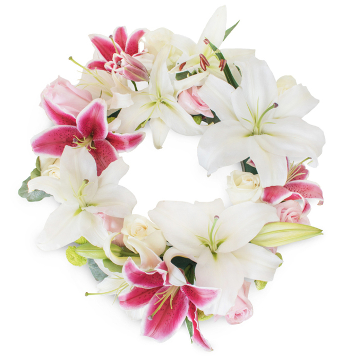 An exquisitely arranged sympathy wreath radiates calm dignity from its perfect circular form, the result of meticulous artistry by local Upper Clapton florists. The dominant star-shaped white lilies unfurl gracefully, each pristine petal curving outward to reveal pale green stamens, setting a foundation of purity reminiscent of the area's bright open spaces. Bespoke fuchsia Stargazer lilies, with their deep pink shades and subtle speckles, infuse energy and focal points throughout the arrangement, contrasting beautifully against the serene whites. Nestled among the lilies, clusters of pale blush and creamy white roses-some buds, some just opening-add a gentle, romantic softness, their ruffled petals offering comfort in times of farewell. Minimalist yet intentional green foliage peeks through, calling to mind the treescape of Hackney Downs, and enhancing the lush abundance of the wreath. The composition's fullness and open center convey continuity and celebration of life, its palette of pale and vivid tones serving both as tribute and gentle comfort. Suspended against a clean white backdrop, this arrangement stands as an elegant, heartfelt offering for memorial services, remembrance, or condolence in the Upper Clapton community, each floral element thoughtfully selected for its beauty and emotional resonance.