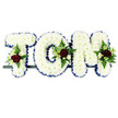 The photograph displays a classic funeral flower name tribute reading TOM, composed of three bold capital letters arranged on a slim support bar against a crisp white background. Each letter is fully covered with snow-white chrysanthemums, their small, tightly packed blooms creating a thick, cushion-like surface that suggests softness and purity. The chrysanthemums' petals are slightly curved and layered, giving the letters a gentle texture and depth that lift them off the supporting frame. Encircling the perimeter of the T, O, and M is a neat border of royal-blue satin ribbon, carefully pleated along the contours to produce a smooth, gleaming edge that defines each letter clearly and adds a traditional, dignified feel. At three key positions - near the top-left of the T, along the right curve of the O, and just above the right side of the M - are tastefully arranged accent sprays of flowers and greenery. Each spray centres on rich, velvety red roses, their petals unfurling in full, rounded blooms. These are complemented by petite white and cream spray roses and slender, elegant white flowers resembling freesia, all nestled into the chrysanthemums. Fresh green foliage and airy fern fronds spill gently around the roses, introducing contrast, movement, and a sense of living garden freshness. With its balanced palette of white, blue, and red, the tribute communicates peace, love, and remembrance in a way that feels both formal and deeply personal. It is the sort of carefully constructed arrangement you might see at a funeral, wake, or memorial service in Upper Clapton, placed beside the coffin or at the front of a chapel to honour Tom's memory with quiet grace.