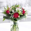 A luxurious red and white bouquet stands proudly in a clear cylindrical glass vase, its neatly trimmed green stems visible through the water like a column of freshness on a bright Upper Clapton morning. At the heart of the arrangement, velvety deep red roses unfurl in layered spirals, each petal rich and saturated, capturing that timeless feeling of romance often shared along the quiet streets off Upper Clapton Road. Nestled closely beside them is a large, deeply ruffled crimson carnation, its intricate, frilled petals adding drama and texture, almost like soft fabric gathered by hand. Around these passionate red blooms, clusters of pure white flowers create a calm, luminous contrast. White alstroemeria open with slender petals marked by fine speckles, while closed white lily buds stretch upward, promising future blossoms and a lingering, elegant fragrance. Light, cloud-like sprays of tiny white gypsophila weave through the design, softening the overall shape and giving an airy, celebratory feel, as if ready for a family gathering near Springfield Park. Lush greenery supports and frames the flowers: broad, glossy dark green leaves curl gracefully around the base, forming a structured collar, while slimmer pointed leaves and eucalyptus-like foliage add depth and movement in varied shades of green. The bouquet rests on a pristine white surface against a softly blurred, bright background, suggesting daylight filtering through a nearby window. Gentle, even lighting highlights every petal and leaf, making the arrangement feel both polished and inviting. This is the sort of romantic, uplifting display a florist in Upper Clapton might recommend for anniversaries, birthdays, or to bring warmth and heartfelt affection into a home or office.