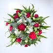 Viewed from directly above against a clean white background, this bouquet appears as a generous, rounded cloud of colour and texture, carefully arranged to feel both modern and romantic. At its centre, deep crimson red roses draw the eye, some still tightly furled and others just beginning to open, their velvety petals curling softly to suggest intimacy and devotion. Around them, ruffled carnations in rich fuchsia and ruby tones add a party-like exuberance, their frilled edges creating a plush, almost fabric-like texture that feels joyful and warm. Nestled between these stronger hues, clusters of small white spray chrysanthemums, like miniature daisies, introduce a fresh, uplifting brightness, each little disk of petals radiating from a golden heart. Threaded delicately through the entire composition are fine sprays of white gypsophila, or baby's breath, so light and airy they look like a soft mist drifting between the blooms. The flowers are framed by an abundance of greenery: broad, pointed dark green leaves, likely from lilies or alstroemeria, provide a bold, architectural framework, while rounded, silvery eucalyptus leaves weave a cooler, blue-green note through the composition, hinting at a gentle, herbal fragrance. The lighting is even and natural, making the reds appear rich but true, the whites crisp, and the greens vivid without harshness. The overall effect is that of a carefully hand-tied bouquet just finished on a florist's bench in Upper Clapton, ready to be carried along the leafy streets near Springfield Park. It feels ideal for marking a birthday, anniversary, or heartfelt thank you, delivering an immediate sense of celebration and affectionate energy as soon as it arrives at the door.