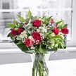A romantic red and white bouquet stands proudly in a clear glass vase on a bright white tabletop, the entire arrangement bathed in gentle daylight from a large, softly blurred window behind. At the heart of the display, velvety crimson red roses-some fully open with layered petals curling outward, others still in elegant bud-create a luxurious focal point. Nestled alongside them are ruffled red carnations in matching tones, their densely packed petals adding volume and a touch of classic charm. Threaded between the richer blooms are crisp white alstroemeria, or Peruvian lilies, some still in bud with pale green-tipped petals, others just beginning to open into graceful, trumpet-like flowers that bring a sense of promise and unfolding joy. Tiny clusters of white gypsophila, or baby's breath, scatter like soft clouds throughout the bouquet, breaking up the stronger colours with a delicate, airy lightness. Around the base and weaving through the design, lush green foliage forms a generous collar: broad, glossy leaves give structure and depth, while slimmer, lighter green stems-reminiscent of eucalyptus or similar greenery-add movement and texture. Through the sides of the vase, the neatly trimmed stems are clearly visible, immersed in clear water that hints at the flowers' freshness, as if just arranged in a local Upper Clapton studio. The neutral, slightly desaturated view beyond the window keeps all attention on the vivid reds, whites, and greens, making the bouquet feel both timeless and contemporary. It evokes the warmth of a heartfelt thank-you, a birthday surprise, or a simple gesture to brighten a quiet home near Springfield Park or along Upper Clapton Road, sending a message of affection and celebration without saying a word.