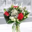 A contemporary mixed bouquet stands in a simple clear glass vase, captured in soft daylight that could be streaming through a townhouse window in Upper Clapton. At its heart, a single coral-red rose and a plush crimson carnation create a vivid focal point, their velvety petals layered and full, suggesting deep emotion and quiet celebration. Around them, clusters of tiny white daisy-like flowers and button chrysanthemums scatter light through the arrangement, their crisp petals forming a halo of purity against the richer tones. Several creamy white lilies, some just beginning to open and others still in smooth green buds, rise elegantly through the centre, adding height and a sense of anticipation, as if the bouquet will keep unfolding over the coming days. The whole design is cradled by broad, glossy emerald leaves, folded and swept in graceful arcs around the base, giving the composition a modern, almost sculptural silhouette. The glass vase reveals clean stems and clear water, reinforcing the impression of freshness, like air after rain over nearby Springfield Park. Light glints off the glass and the varnished foliage, creating subtle reflections that echo the bouquet's clean fragrance-leafy, slightly sweet, and calming. The mood is uplifting yet composed, perfect for a birthday breakfast table, a new home near Clapton Common, or a simple thank-you delivered quietly to a friend. Every detail, from the careful spacing of the blooms to the balance of red, white, and green, speaks of a florist's steady hand and an intention to bring a gentle sense of joy and renewal into an everyday London room.