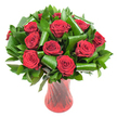 A lush, rounded bouquet of a dozen or more deep crimson roses sits like a jewel in this image, each bloom fully open with velvety, spiralled petals that catch the light and read as almost wine-dark at their centers. The roses are densely clustered to form a generous dome, their smooth, satiny surfaces contrasted by an abundance of glossy dark green foliage-small Ruscus- or Salal-like leaves that create a rich, verdant backdrop and cradle each head. Larger, bright emerald leaves are artfully folded and rolled into cylindrical loops and dramatic spirals, their glossy edges forming architectural arcs that give the classic rose presentation a contemporary, sculptural rhythm. The whole composition is anchored in a tall, slender translucent ruby-red glass vase with a subtle twisted, ribbed texture; vertical lines on the vase catch and reflect the soft, even studio lighting, echoing the roses' deep red and adding a quiet sense of movement. Set against a clean white background, the saturated reds and greens pop with clarity, allowing an eye-and a hand guided by memory-to trace petal by petal, leaf by leaf. The arrangement exudes classic romance and considered craftsmanship, its scent suggested as a soft, warm note that lingers like the memory of a special evening. As a local touch, this style of bouquet is a familiar sight at intimate gatherings and anniversary dinners around Clapton Common and the quieter streets off Lea Bridge Road, where floral gifts often mark milestones with the same timeless passion and thoughtful design.