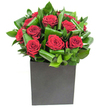 In this elegantly modern composition, a cluster of rich, velvety red roses forms the immediate focal point, each bloom unfolding in lush, spiral layers that seem to glow against the clean lighting. The petals are saturated crimson, their soft, almost plush texture contrasted by bands of glossy green foliage that have been thoughtfully curled and looped to create a rhythmic, sculptural frame. Those broad leaves catch the light at their creases, producing subtle highlights and a faint sheen that reinforces the arrangement's sense of movement and depth. The flowers sit low and secure in a matte black, square box vase whose geometric lines lend the display a contemporary edge and anchor the composition with quiet confidence. The scene is lit with clear, natural-feeling light-crisp enough to reveal the velvet surface of each petal and the waxy translucence of the foliage-so the arrangement reads as both intimate and distinctly modern. From a sensory perspective, the dominant impression is of classic rose perfume softened by green, leafy notes, the scent clean and uplifting rather than heavy. As a florist I picture this design placed on a city table or reception desk near Clapton Common or Chatsworth Road market, a statement that reads as both romantic and composed, ideal for anniversaries, meaningful apologies, or elevating an everyday corner into something thoughtful. The craftsmanship-careful spacing, consistent spiral form, and the tactile contrast between petals and leaves-speaks to considered making and a desire to communicate affection without excess, offering a bouquet that feels at once bold and quietly personal.